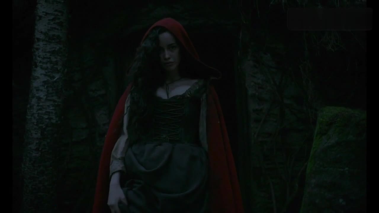 Sarah Stephens in a red hood at the doorway of a hut in The Witch